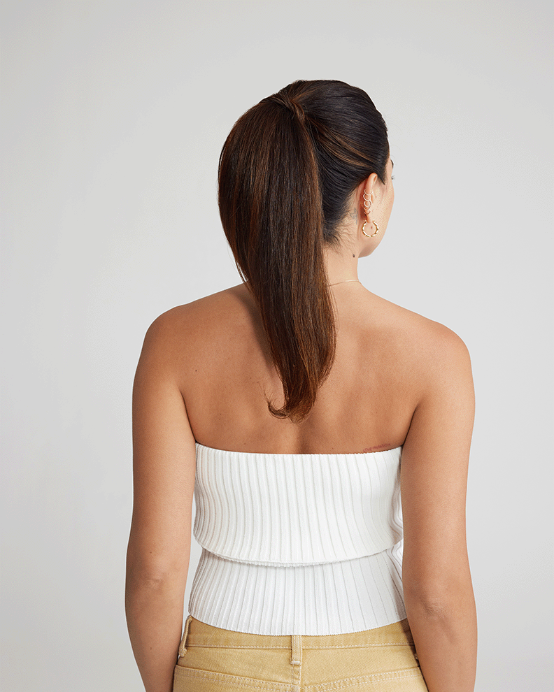 A woman in a white top and tan pants, viewed from the back, preparing to apply hair extensions.
