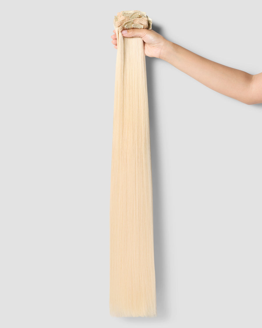 A hand delicately grips a long, clip-in beachy blonde hair extension, highlighting its length and style.