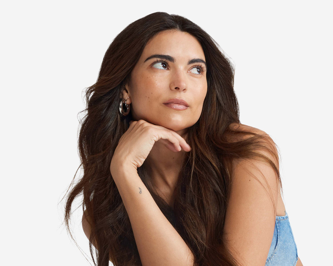 A woman with long wavy dark brown hair wearing hoop earrings and a denim top, resting her chin on her hand and looking upward