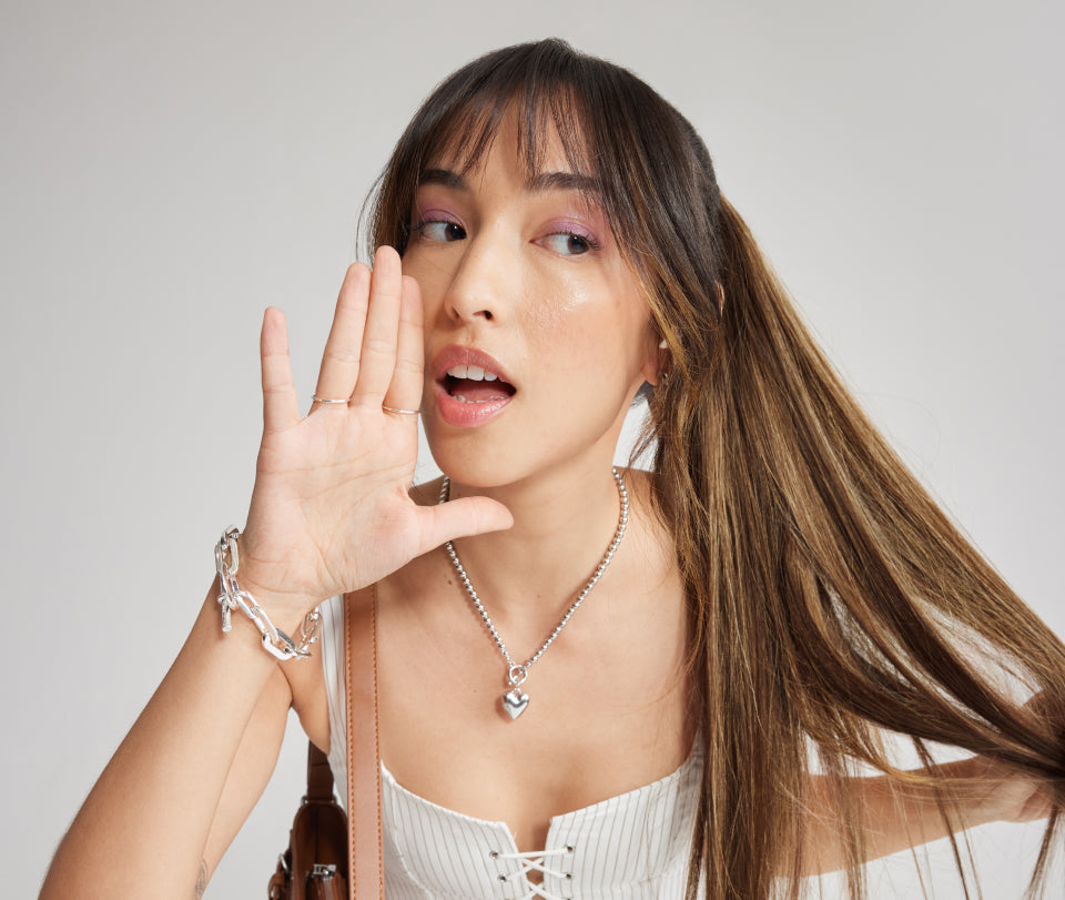 A woman with long, straight brown hair styled in a high ponytail, wearing a white lace-up top and silver jewelry.