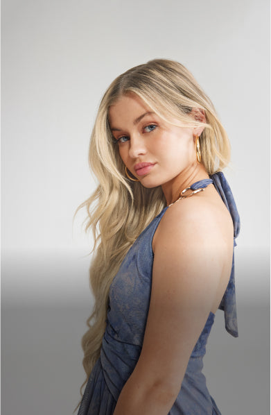 A blonde woman wearing a blue dress, showcasing her long hair extensions curled posting to the side.