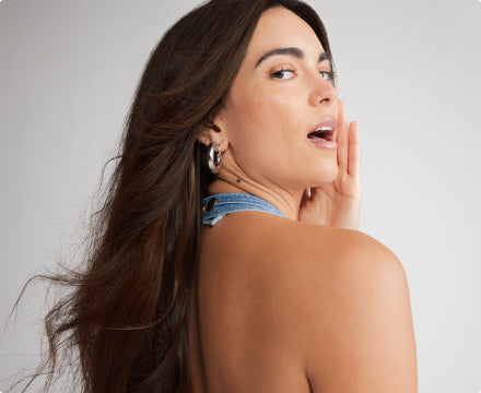 Woman with long brown Zala hair extensions in a denim halter top poses smiling with hand near her face.