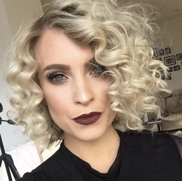 A close-up of a woman with short, platinum blonde, tightly defined curls and bold makeup, including dark burgundy lipstick.
