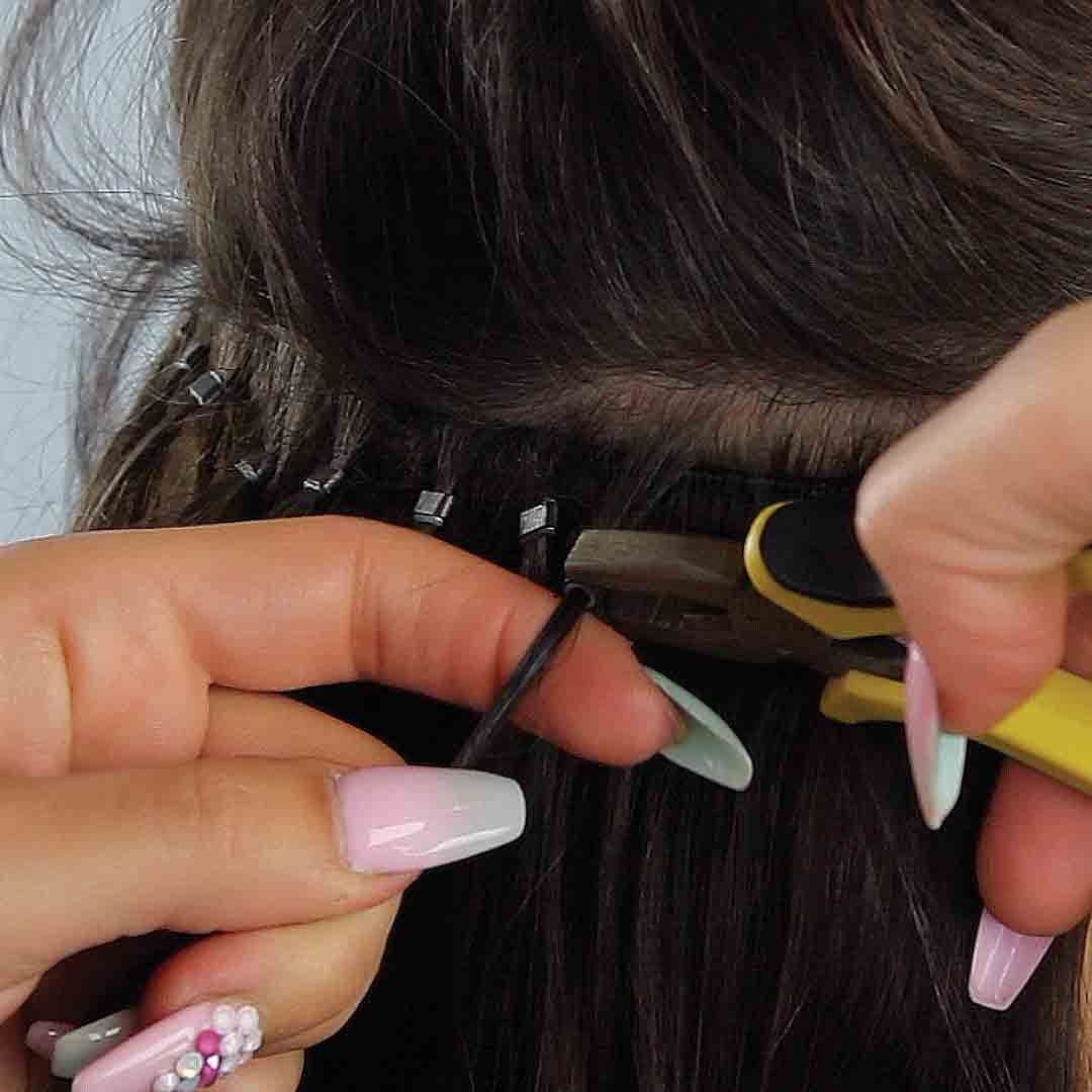 A close-up of hair extension installation using microbeads and pliers on dark brown natural hair.