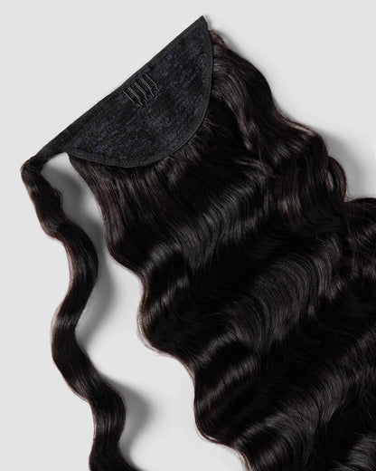 A hand gently holds a long, curly ponytail of natural black hair extensions, showcasing its rich brown hue.